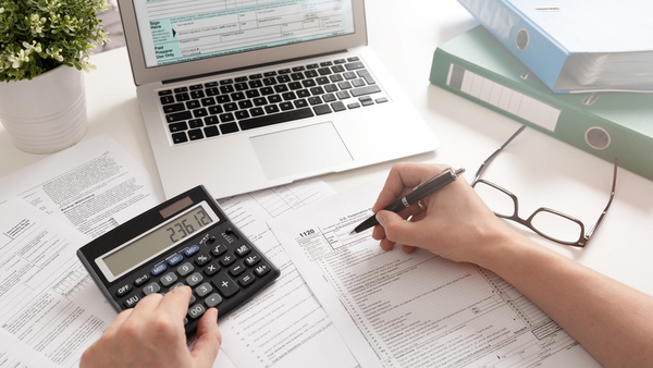 Hands with calculator, laptop, forms on table, pen, and binders