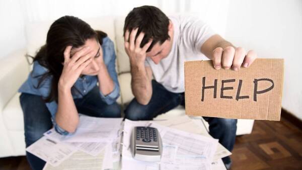 Man holding up help sign with woman with her head in hands. A calculator and bills lay on the  table